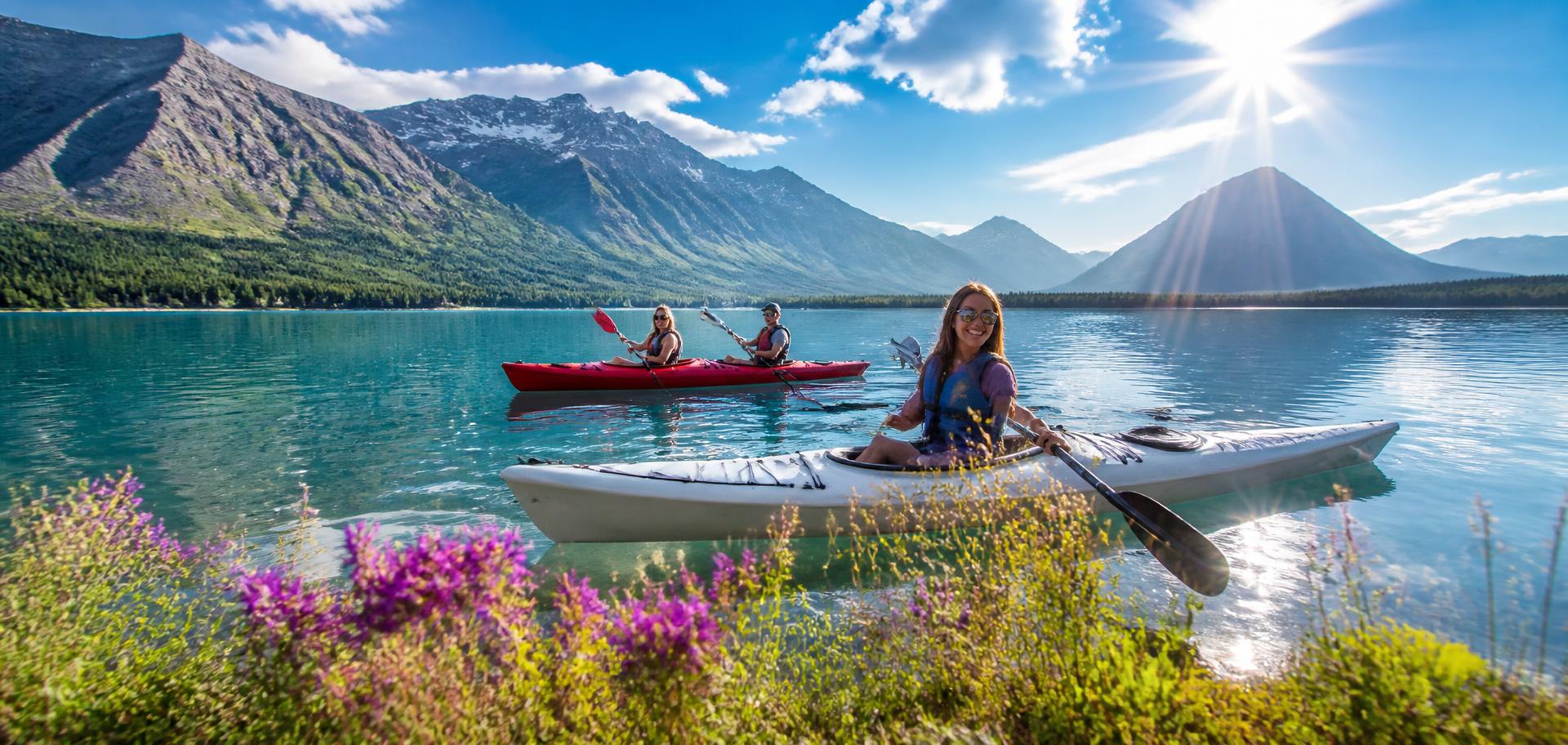 Friends kayaking on a bright blue Alberta mountain lake with sun flare and wildflowers